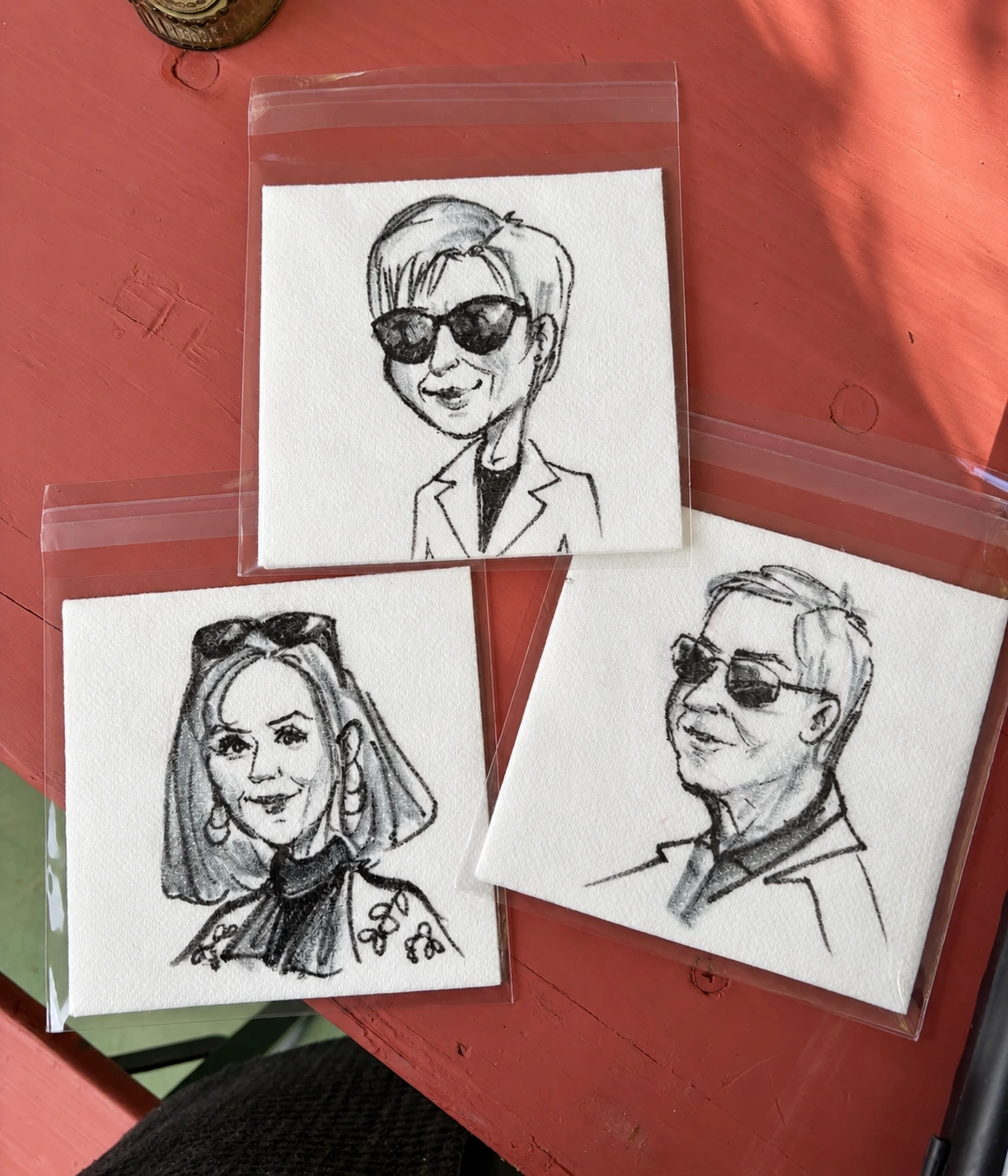 Three hand-drawn portrait napkins in clear sleeves on a coral table