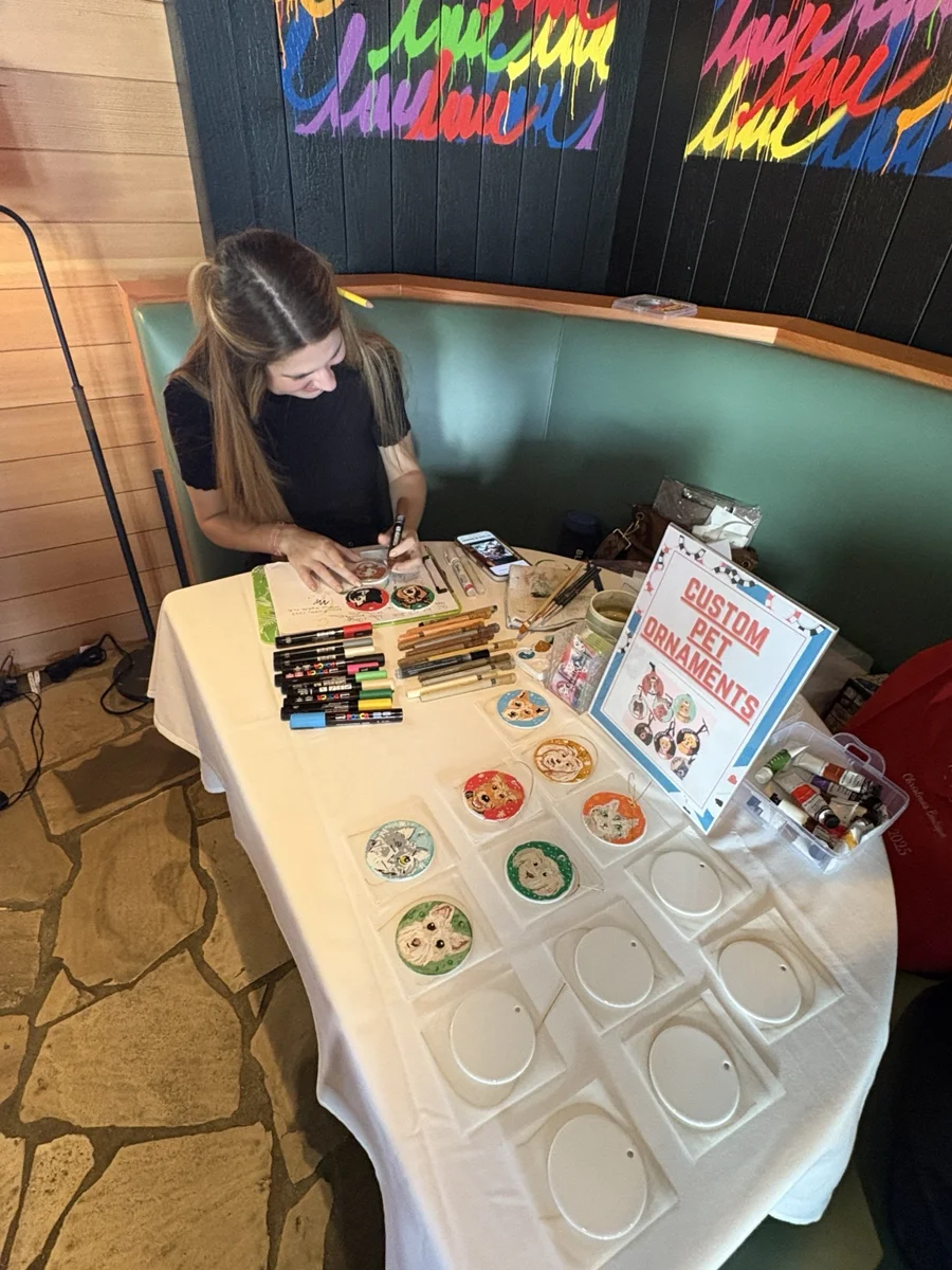 CreateLine artist painting pet ornaments at an event, with finished examples on the table