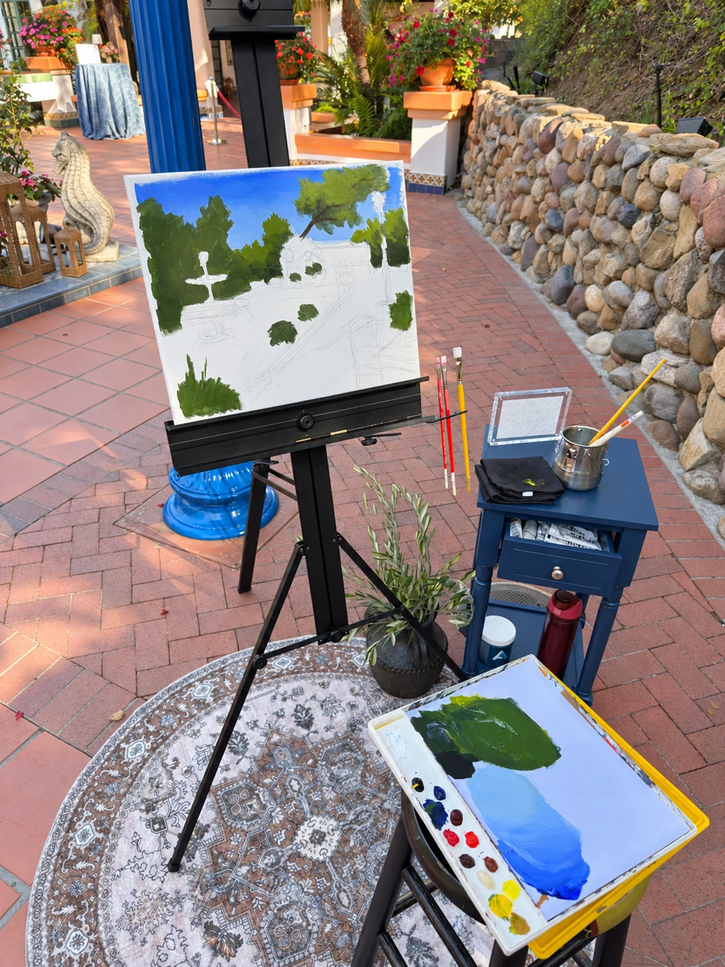 A courtyard painting in progress on an easel beside an open paint palette